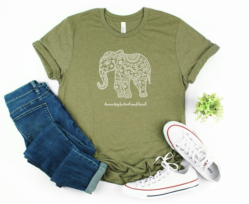 May include: An olive green short-sleeve t-shirt featuring a white graphic of an elephant filled with intricate floral patterns and the text "dream big be kind work hard" printed below the elephant, paired with blue denim jeans and white sneakers.
