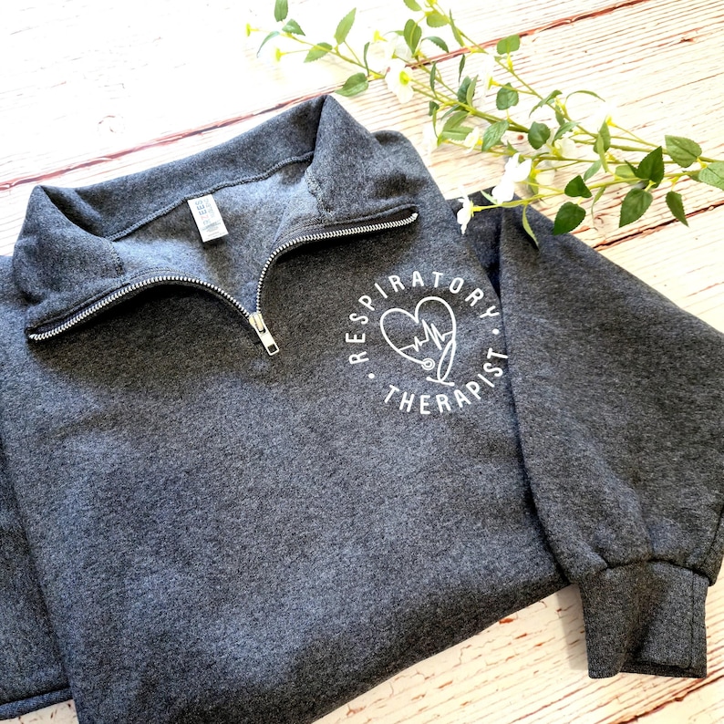 May include: A dark gray sweatshirt with a quarter-zip neckline. The sweatshirt features a white circular graphic with a heart, stethoscope, and the words "Respiratory Therapist". The sleeves are long and the material appears soft.