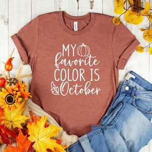 May include: A rust-colored t-shirt with rolled-up sleeves. The shirt features the white text "MY favorite COLOR IS October" with a pumpkin and leaf graphic. The shirt is paired with blue jeans and autumn-themed decorations.