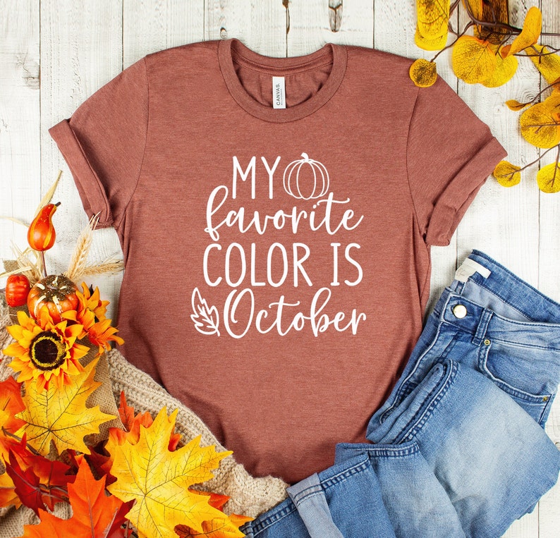 May include: A rust-colored t-shirt with rolled-up sleeves. The shirt features the white text "MY favorite COLOR IS October" with a pumpkin and leaf graphic. The shirt is paired with blue jeans and autumn-themed decorations.