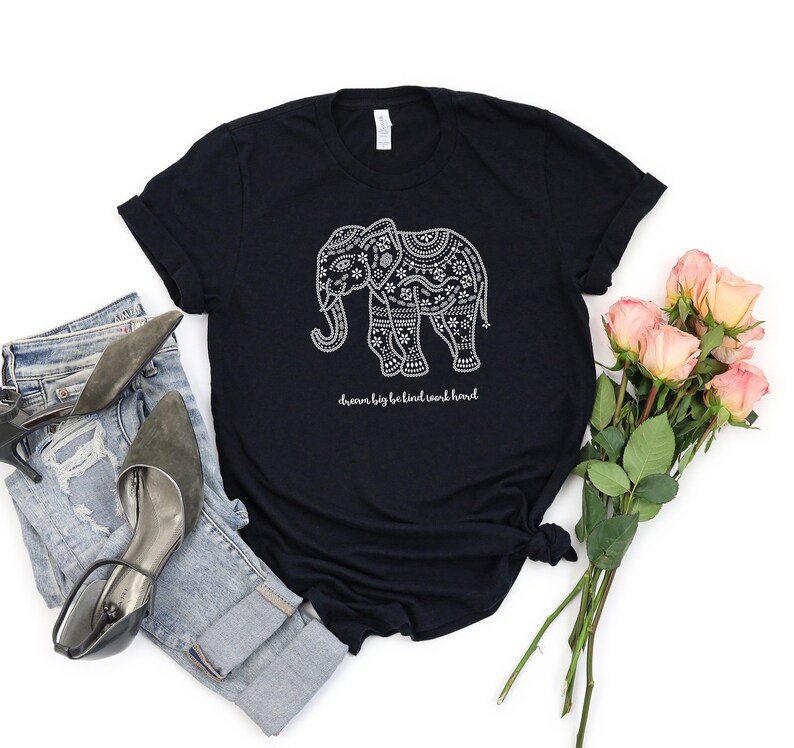 May include: A black short-sleeved t-shirt featuring a white graphic of an elephant decorated with intricate floral and geometric patterns. Below the elephant, the text reads "dream big be kind work hard" in a white cursive script font.