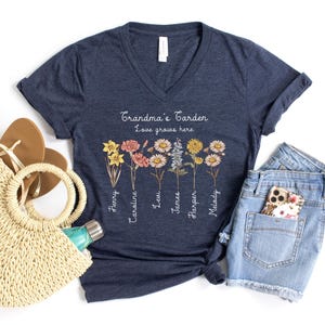 May include: Navy blue V-neck t-shirt with the text "Grandma's Garden Love grows here." The shirt features a floral design with names below each flower. A straw tote bag, sandals, and denim shorts are also in the image.