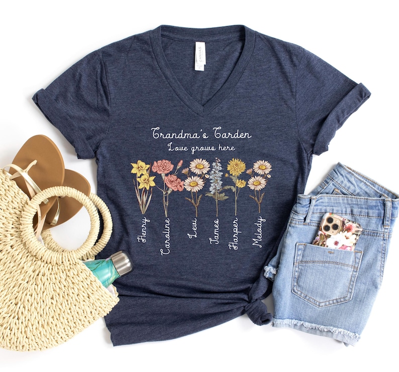 May include: Navy blue V-neck t-shirt with the text "Grandma's Garden Love grows here." The shirt features a floral design with names below each flower. A straw tote bag, sandals, and denim shorts are also in the image.