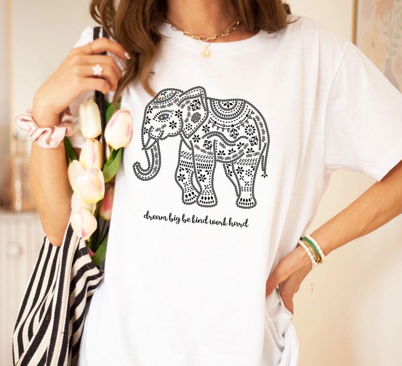 May include: A white short-sleeve t-shirt featuring a black graphic of an elephant decorated with intricate patterns. Below the elephant, the text reads, dream big be kind work hard. The design is printed in black ink on the front of the shirt.
