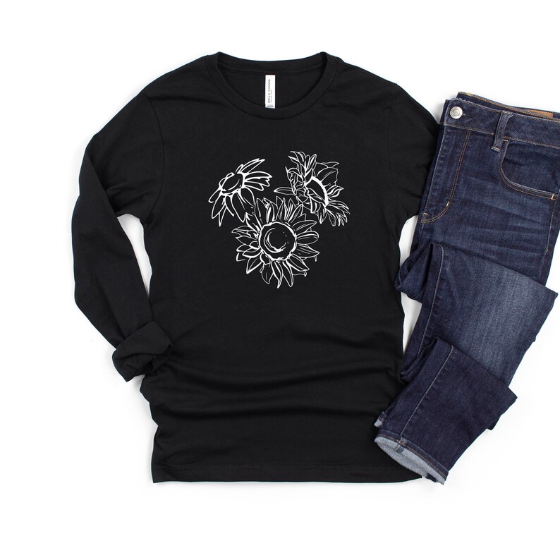 May include: Black long-sleeve shirt with a white sunflower graphic. The graphic features three sunflowers in a sketched style. The shirt is paired with dark wash blue jeans. The shirt is made of a soft, comfortable material.