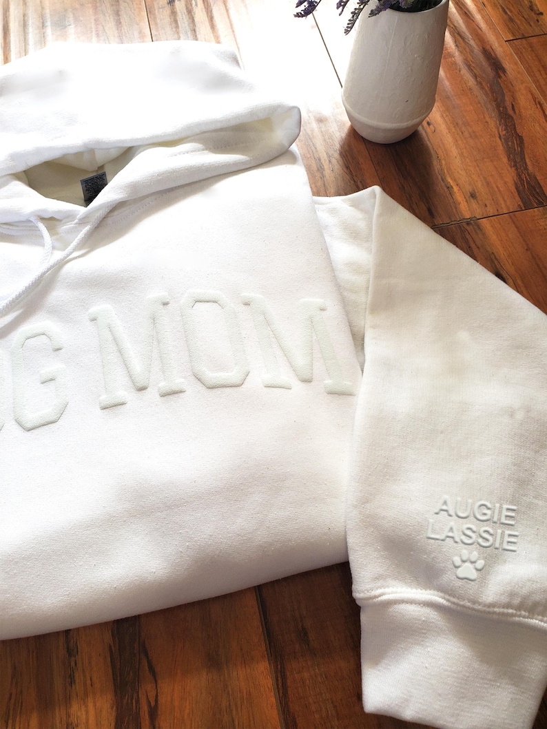 May include: A white hoodie with the words "DOG MOM" embossed on the front. The sleeve features the name "AUGIE LASSIE" and a paw print. The hoodie is made of a soft material and has a hood with a drawstring.