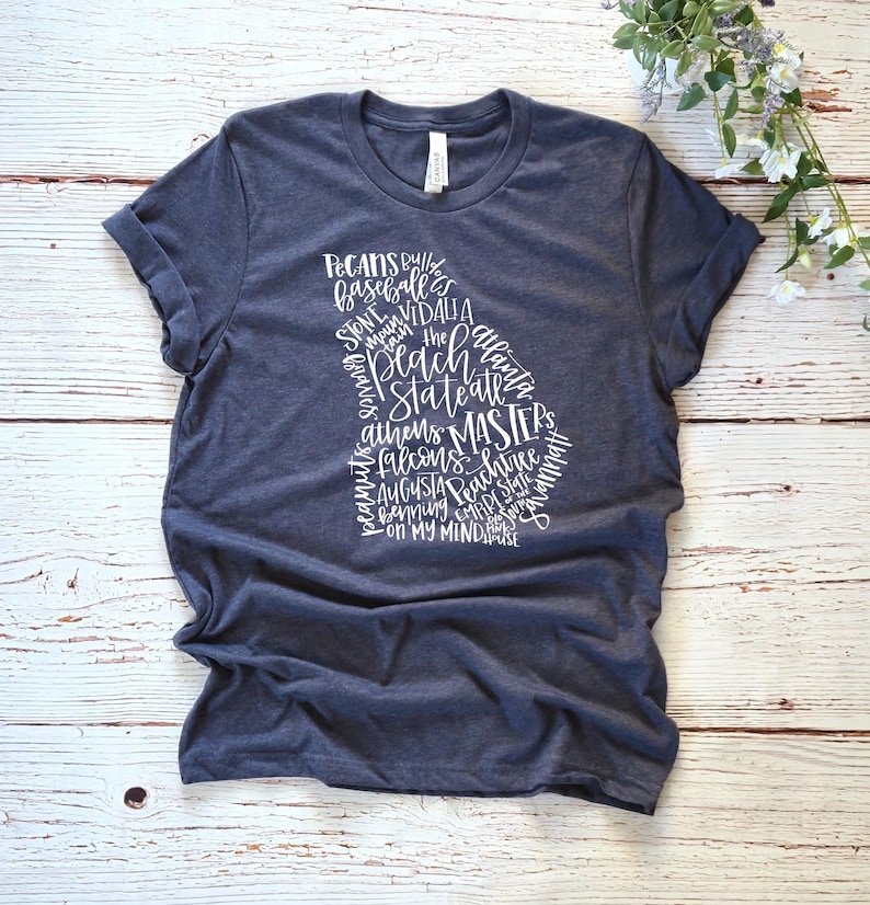 May include: A navy blue t-shirt featuring a white graphic of the state of Georgia, filled with words like "Peaches," "Atlanta," and "Masters." The shirt is made of soft material and has rolled-up sleeves.