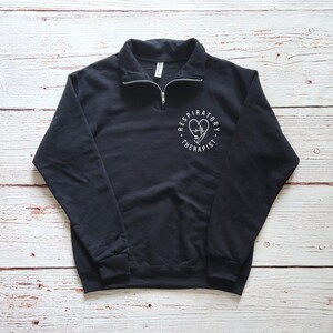 May include: Black quarter-zip sweatshirt with a white graphic on the left chest. The graphic features a heart, stethoscope, and the words "Respiratory Therapist" in a circular design. The sweatshirt has a ribbed collar and cuffs.