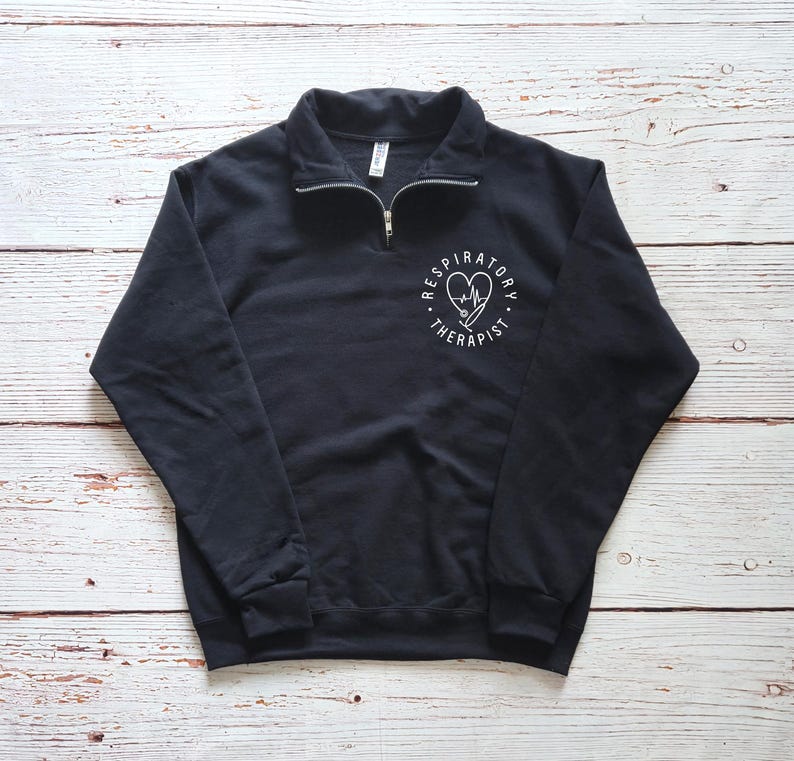 May include: Black quarter-zip sweatshirt with a white graphic on the left chest. The graphic features a heart, stethoscope, and the words "Respiratory Therapist" in a circular design. The sweatshirt has a ribbed collar and cuffs.