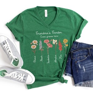 May include: A green V-neck t-shirt with the text "Grandma's Garden Love grows here" and floral illustrations. The names Olivia, Noah, Isabella, Amelia, and Mia are printed below the flowers. Gray sneakers and denim shorts are also in the image.