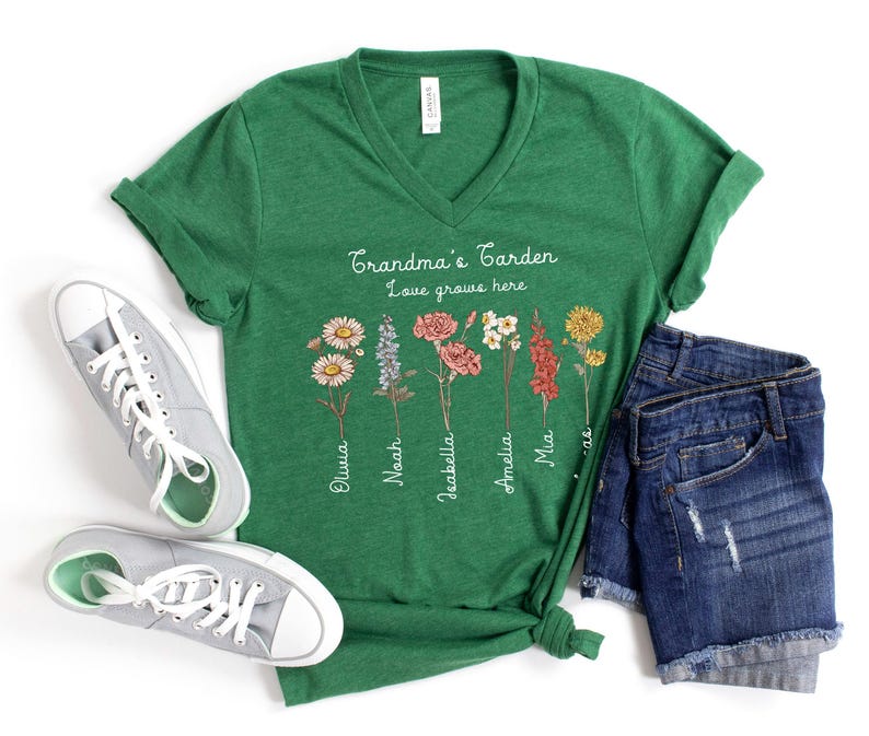 May include: A green V-neck t-shirt with the text "Grandma's Garden Love grows here" and floral illustrations. The names Olivia, Noah, Isabella, Amelia, and Mia are printed below the flowers. Gray sneakers and denim shorts are also in the image.