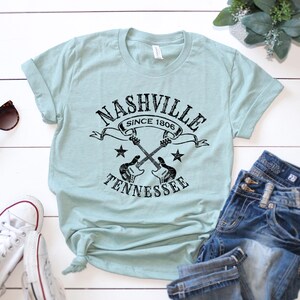 May include: A light blue t-shirt with a black graphic that reads "Nashville Since 1806 Tennessee" with crossed guitars and stars. The shirt is paired with blue jeans and white sneakers.