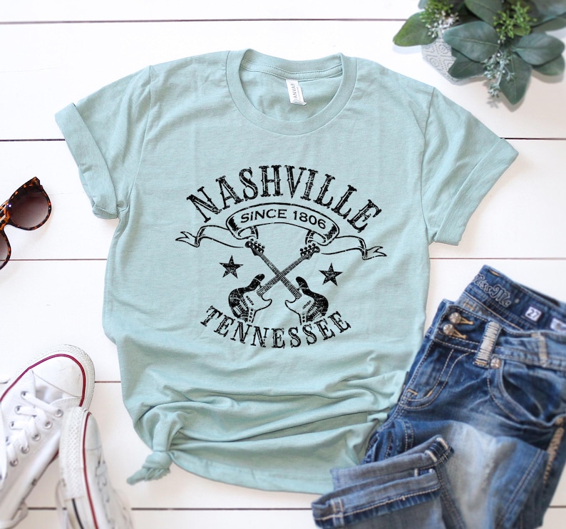 May include: A light blue t-shirt with a black graphic that reads "Nashville Since 1806 Tennessee" with crossed guitars and stars. The shirt is paired with blue jeans and white sneakers.
