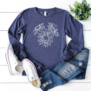 May include: A navy blue long-sleeve shirt with a white sunflower graphic. The shirt is paired with blue jeans and white sneakers. A small green plant is in the background.