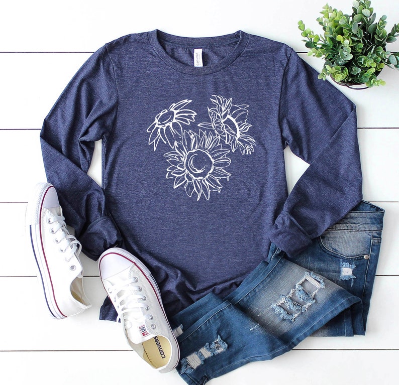 May include: A navy blue long-sleeve shirt with a white sunflower graphic. The shirt is paired with blue jeans and white sneakers. A small green plant is in the background.