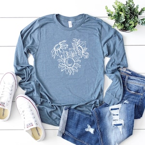May include: A blue long-sleeve shirt with a white floral design, paired with blue jeans and white sneakers. The shirt features a sunflower and other flower outlines. The jeans are distressed. A small potted plant is also visible.