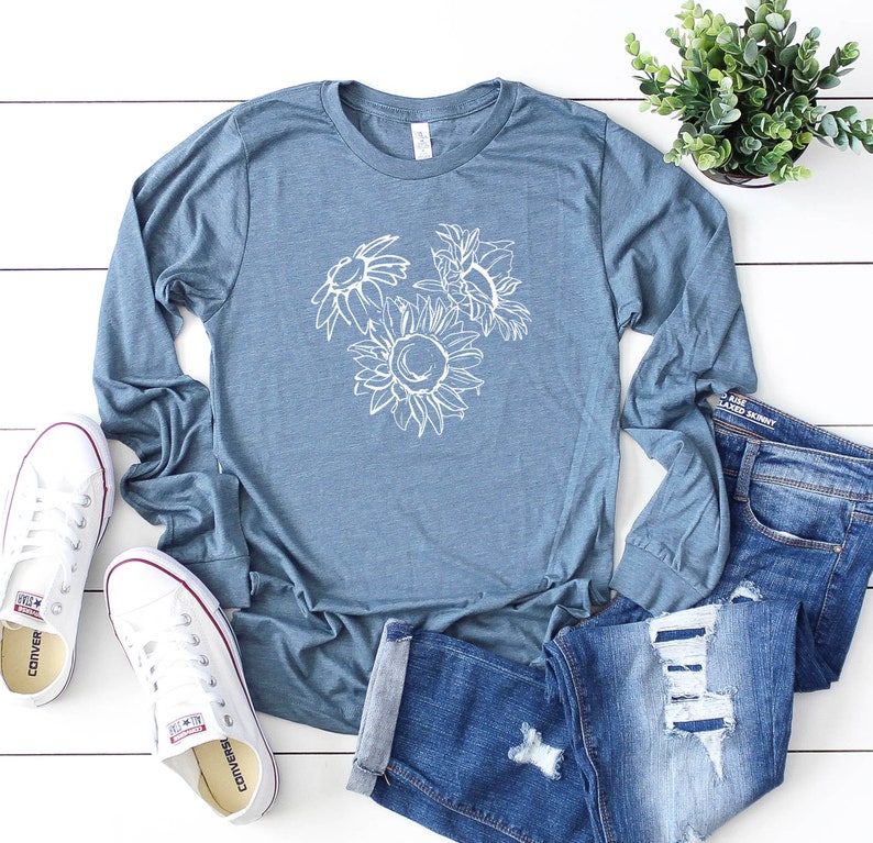 May include: A blue long-sleeve shirt with a white floral design, paired with blue jeans and white sneakers. The shirt features a sunflower and other flower outlines. The jeans are distressed. A small potted plant is also visible.