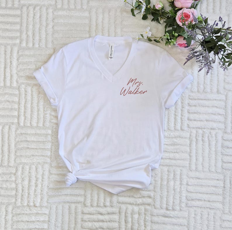 Op de afbeelding: Een witte V-hals T-shirt met opgerolde mouwen. De tekst "Mrs. Walker" is in cursieve letters op de linkerborst gedrukt. Het shirt is gemaakt van een zacht materiaal en is geschikt voor dagelijks gebruik.