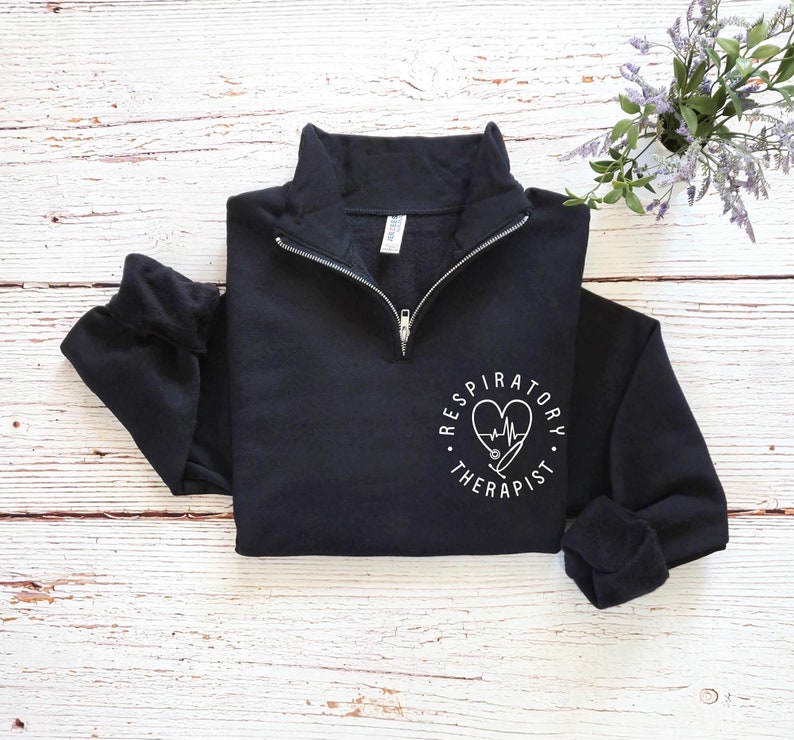 May include: Black quarter-zip sweatshirt with white text and a heart-shaped design. The design includes a stethoscope and the words "Respiratory Therapist" in a circular pattern. The sweatshirt has a high collar and long sleeves.