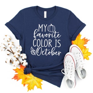 May include: Navy blue t-shirt with the text "MY favorite COLOR IS October" in white. The shirt is decorated with a pumpkin and leaf graphic. White sneakers and fall leaves are arranged around the shirt.