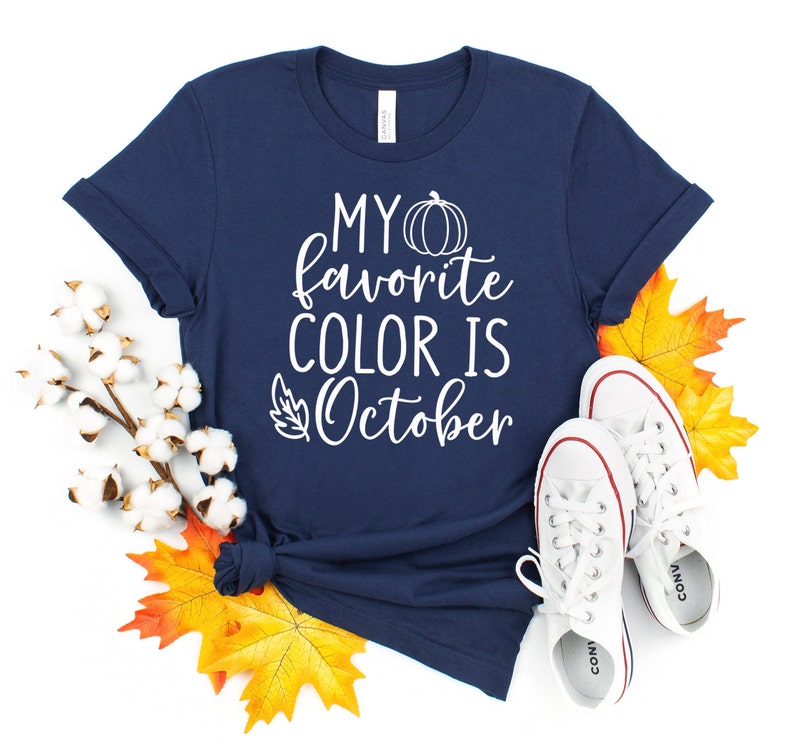 May include: Navy blue t-shirt with the text "MY favorite COLOR IS October" in white. The shirt is decorated with a pumpkin and leaf graphic. White sneakers and fall leaves are arranged around the shirt.