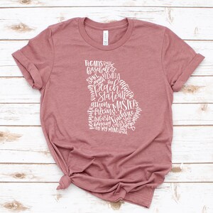 May include: A dusty rose t-shirt featuring a white graphic in the shape of the state of Georgia. The graphic is composed of various words, including "Peach State" and city names like "Atlanta" and "Augusta". The shirt is knotted at the waist.