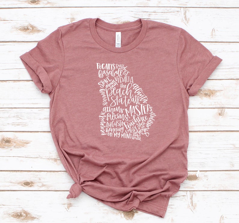 May include: A dusty rose t-shirt featuring a white graphic in the shape of the state of Georgia. The graphic is composed of various words, including "Peach State" and city names like "Atlanta" and "Augusta". The shirt is knotted at the waist.