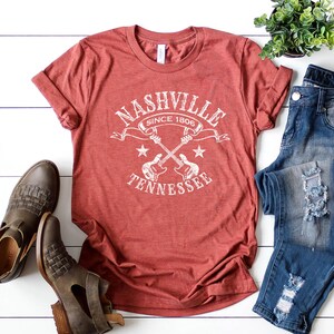 May include: A red t-shirt with the text "Nashville Tennessee Since 1806" in white, featuring crossed guitars and stars. The shirt is paired with blue jeans and brown leather boots, creating a casual, country-inspired ensemble.