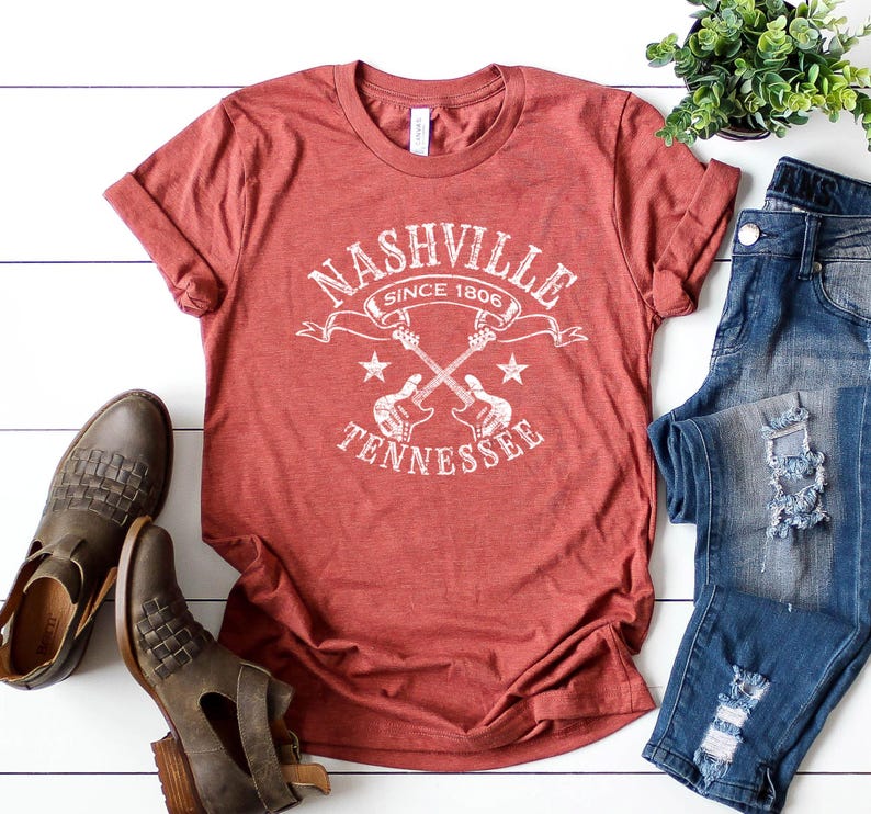 May include: A red t-shirt with the text "Nashville Tennessee Since 1806" in white, featuring crossed guitars and stars. The shirt is paired with blue jeans and brown leather boots, creating a casual, country-inspired ensemble.