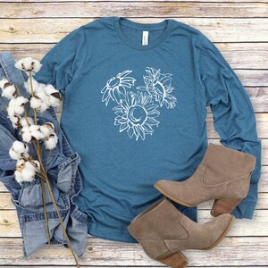 May include: Teal long-sleeve shirt with a white sunflower graphic. The shirt is paired with denim jeans, cotton stems, and brown suede ankle boots with a wooden heel. The items are arranged on a light wood-grain surface.