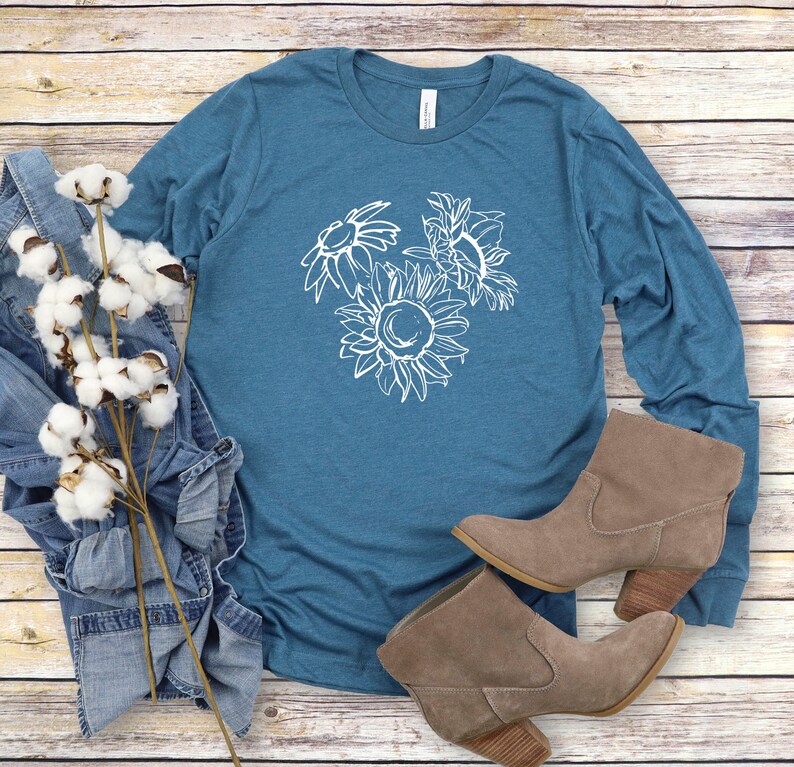 May include: Teal long-sleeve shirt with a white sunflower graphic. The shirt is paired with denim jeans, cotton stems, and brown suede ankle boots with a wooden heel. The items are arranged on a light wood-grain surface.