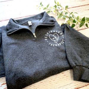 May include: A charcoal gray sweatshirt with a quarter-zip neckline. The sweatshirt features the text "Respiratory Therapist" in a white circular design with a heart and stethoscope graphic. The sleeves have a ribbed cuff.