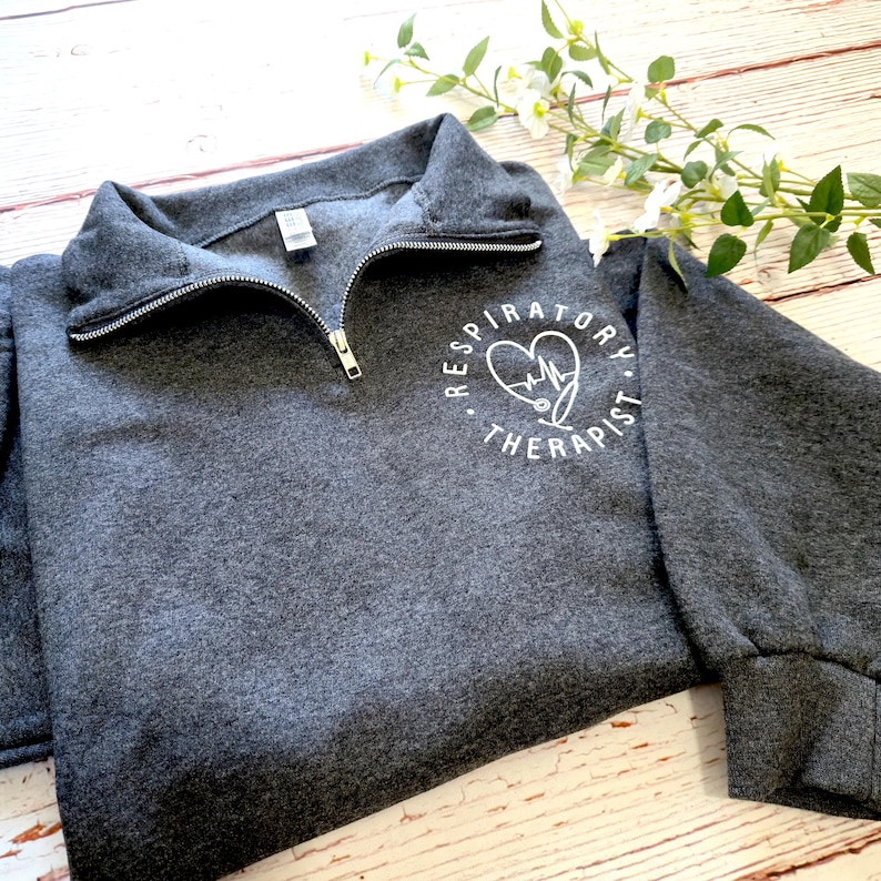 May include: A charcoal gray sweatshirt with a quarter-zip neckline. The sweatshirt features the text "Respiratory Therapist" in a white circular design with a heart and stethoscope graphic. The sleeves have a ribbed cuff.