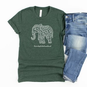 May include: A dark green heathered t-shirt featuring a white graphic of an elephant decorated with intricate patterns and the text "dream big be kind work hard" positioned below the elephant, paired with a pair of blue denim jeans.