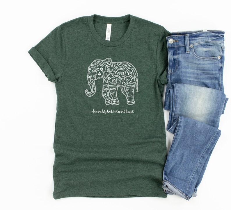 May include: A dark green heathered t-shirt featuring a white graphic of an elephant decorated with intricate patterns and the text "dream big be kind work hard" positioned below the elephant, paired with a pair of blue denim jeans.