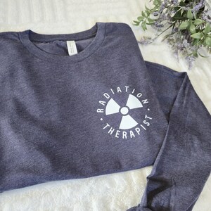 以下が含まれることがあります： 白のグラフィックが特徴の、ネイビーブルーの長袖シャツ。「RADIATION THERAPIST」の文字が放射線マークを囲んでいます。柔らかい素材で作られたシャツです。
