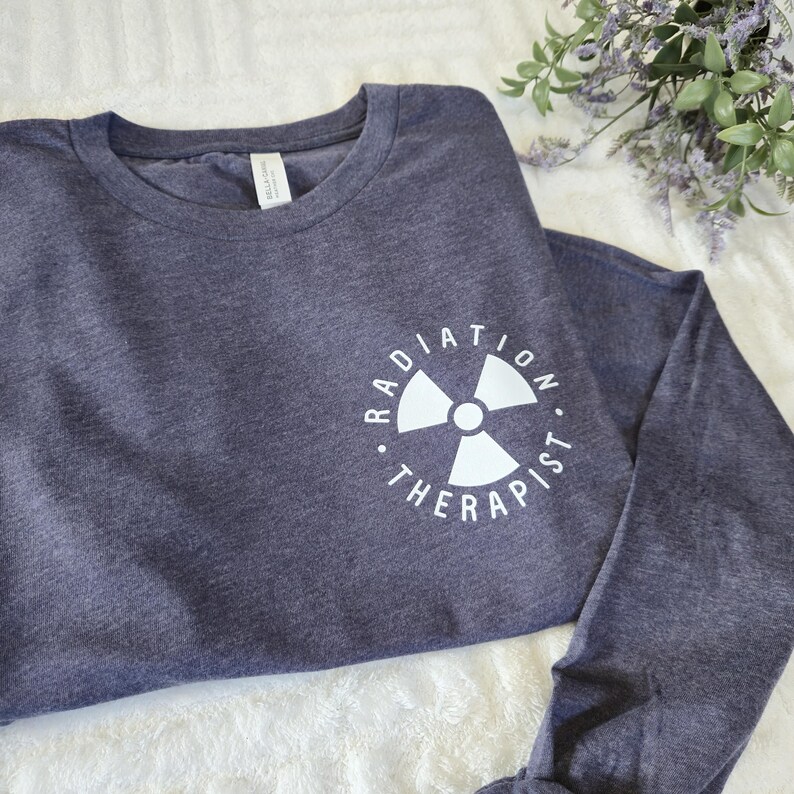 以下が含まれることがあります： 白のグラフィックが特徴の、ネイビーブルーの長袖シャツ。「RADIATION THERAPIST」の文字が放射線マークを囲んでいます。柔らかい素材で作られたシャツです。