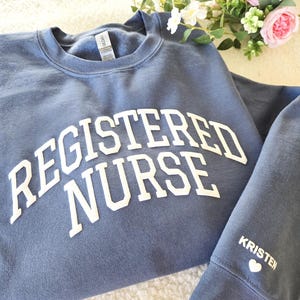 May include: A dusty blue crewneck sweatshirt featuring the white text REGISTERED NURSE in an arched font across the chest. The sleeve displays the name KRISTEN in white capital letters above a small white heart icon, with a Gildan brand label.