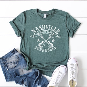 May include: Green t-shirt with a white graphic that reads "Nashville Tennessee Since 1806" with crossed guitars and stars. The shirt is paired with blue jeans and white sneakers.