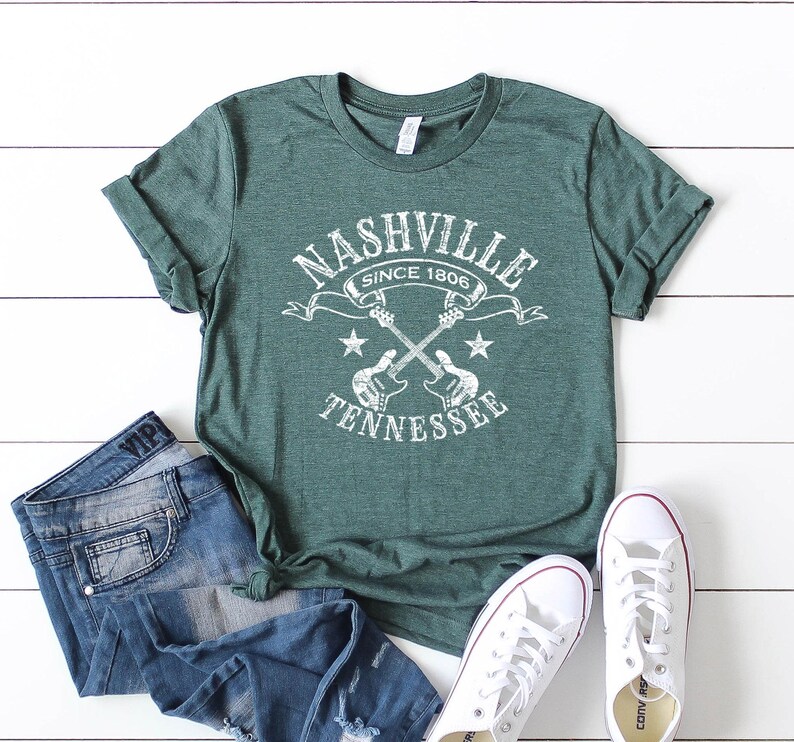 May include: Green t-shirt with a white graphic that reads "Nashville Tennessee Since 1806" with crossed guitars and stars. The shirt is paired with blue jeans and white sneakers.