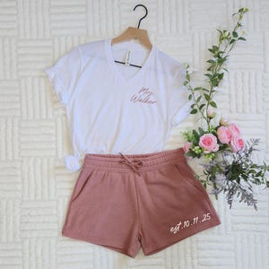Op de afbeelding: Een witte V-hals t-shirt met "Mrs. Walker" in sierlijke letters, gecombineerd met oudroze shorts. De shorts hebben een taille met trekkoord en de tekst "est.10.11.25". De set wordt gepresenteerd met bloemen.