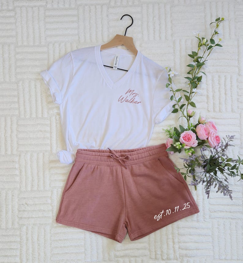 Op de afbeelding: Een witte V-hals t-shirt met "Mrs. Walker" in sierlijke letters, gecombineerd met oudroze shorts. De shorts hebben een taille met trekkoord en de tekst "est.10.11.25". De set wordt gepresenteerd met bloemen.