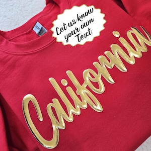 May include: A red sweatshirt with the word "California" in gold lettering. A white and gold text bubble at the top says "Let us know your own Text". The sweatshirt has a crew neck and long sleeves.