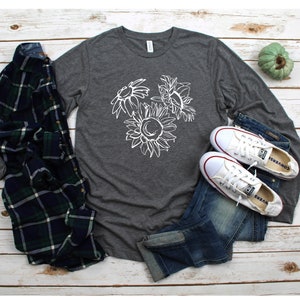 May include: A dark gray long-sleeve shirt with a white sunflower graphic, paired with blue jeans, white sneakers, and a green, blue, and black plaid shirt. The shirt and jeans are laid on a light wood surface.