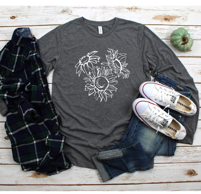 May include: A dark gray long-sleeve shirt with a white sunflower graphic, paired with blue jeans, white sneakers, and a green, blue, and black plaid shirt. The shirt and jeans are laid on a light wood surface.