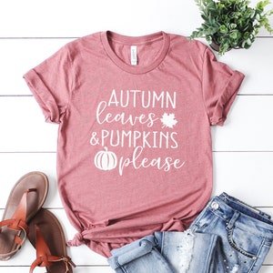 May include: A dusty rose t-shirt with the words "AUTUMN leaves & PUMPKINS please" in white script. A white maple leaf and pumpkin graphic are included. The shirt is paired with blue jeans and brown sandals, suggesting a fall theme.