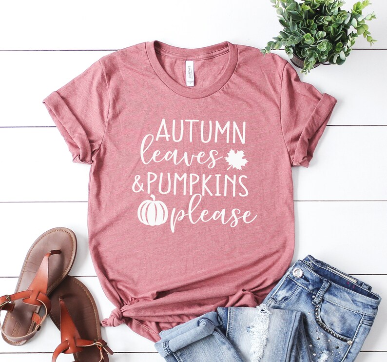 May include: A dusty rose t-shirt with the words "AUTUMN leaves & PUMPKINS please" in white script. A white maple leaf and pumpkin graphic are included. The shirt is paired with blue jeans and brown sandals, suggesting a fall theme.
