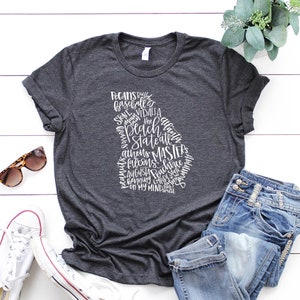 May include: Dark gray t-shirt featuring a white outline of the state of Georgia filled with text. The text includes words like "Pecans", "Braves", "Athens", and "Augusta". The shirt is paired with blue jeans, sunglasses, and white sneakers.