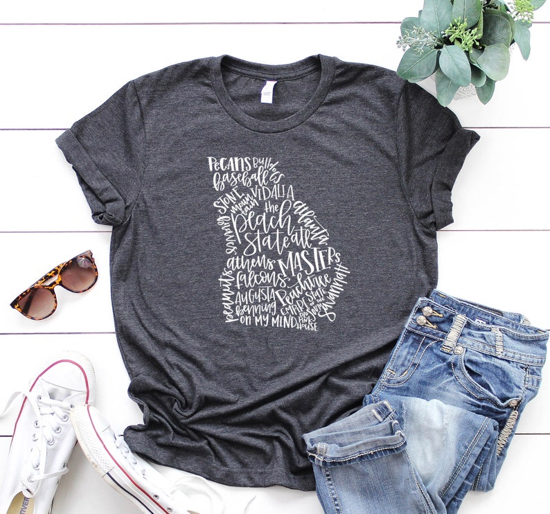 May include: Dark gray t-shirt featuring a white outline of the state of Georgia filled with text. The text includes words like "Pecans", "Braves", "Athens", and "Augusta". The shirt is paired with blue jeans, sunglasses, and white sneakers.