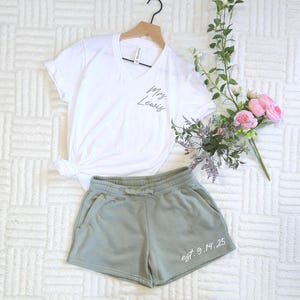 May include: A white V-neck t-shirt with "Mrs. Lewis" printed on it, paired with sage green shorts. The shorts have "est. 9.14.25" printed on them. The ensemble is displayed with a bouquet of pink roses and greenery.